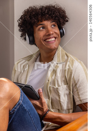 African american man sitting on wooden bench in room corner using tablet with headphones African american man sitting on wooden bench in room corner using tablet with headphones 140564306