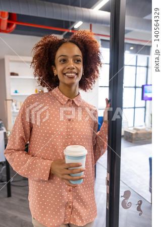 African american woman leaning against glass door in office by white shelving with coffee cup 140564329