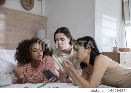 Diverse female friends leaning on bed in bedroom focusing smartphone with green pencil papers Diverse female friends leaning on bed in bedroom focusing smartphone with green pencil papers 140564374