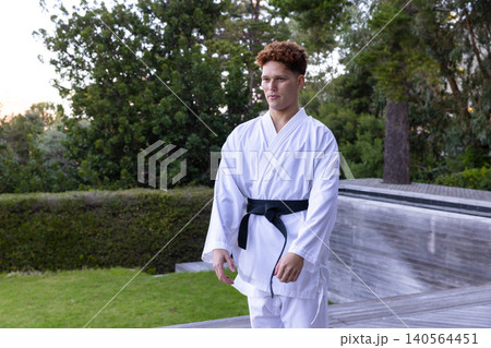 Male karate student practicing on deck by trimmed hedge wearing white gi and black belt Male karate student practicing on deck by trimmed hedge wearing white gi and black belt 140564451