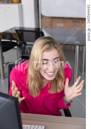 Woman in pink blouse leaning forward at desk in office raising hands with computer setup 140564491