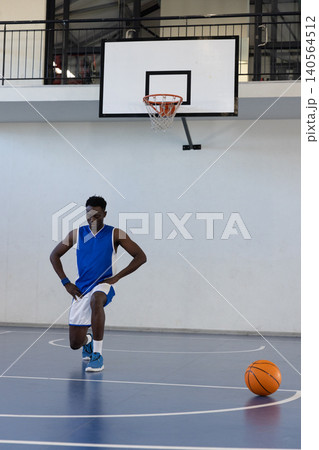 Kneeling african american male in blue jersey stretching on court near hoop with basketball Kneeling african american male in blue jersey stretching on court near hoop with basketball 140564512