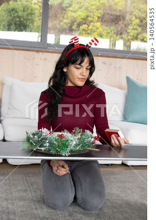 Woman kneeling at home arranging christmas wreath centerpiece on coffee table holding gift box Woman kneeling at home arranging christmas wreath centerpiece on coffee table holding gift box 140564535