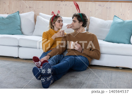 Couple sitting at home wearing antler headbands and knitted socks holding ceramic mugs by wood wall Couple sitting at home wearing antler headbands and knitted socks holding ceramic mugs by wood wall 140564537