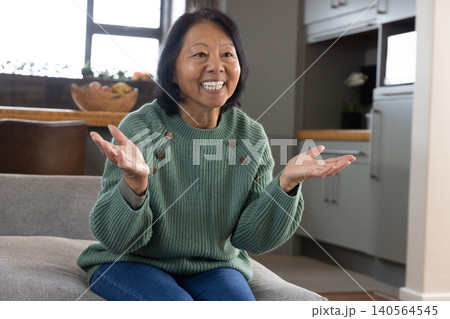 Asian woman wearing green sweater gesturing on gray sofa in living room with fruit bowl Asian woman wearing green sweater gesturing on gray sofa in living room with fruit bowl 140564545