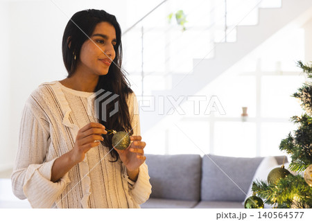 Indian woman holding green Christmas ornament and placing on tree in living room, copy space 140564577