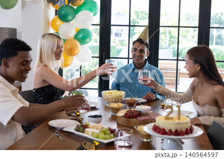 Diverse friends wearing party hats around wooden table celebrating birthday with cake, balloons Diverse friends wearing party hats around wooden table celebrating birthday with cake, balloons 140564597