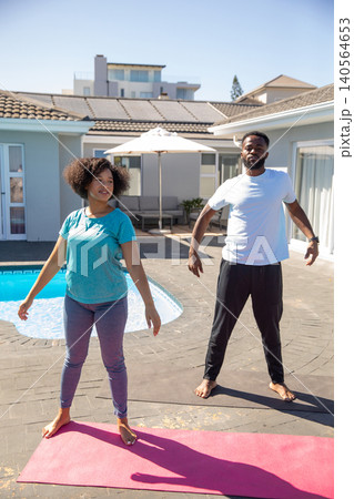 Diverse couple wearing sportswear practicing stretching exercises on yoga mats, backyard pool Diverse couple wearing sportswear practicing stretching exercises on yoga mats, backyard pool 140564653