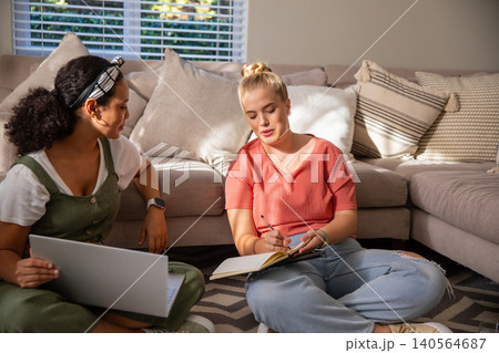 Diverse female friends studying on patterned rug in living room with laptop, notebook Diverse female friends studying on patterned rug in living room with laptop, notebook 140564687