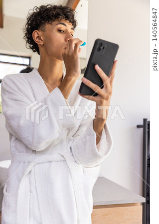 Man in white bathrobe checking smartphone, brushing teeth with blue toothbrush at vanity 140564847