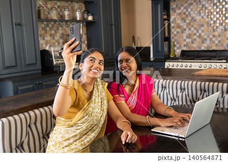Indian mother and daughter taking selfie with smartphone while using laptop at kitchen table Indian mother and daughter taking selfie with smartphone while using laptop at kitchen table 140564871