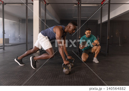 Diverse male trainer and trainee monitoring tablet timer and performing kettlebell push-ups at gym Diverse male trainer and trainee monitoring tablet timer and performing kettlebell push-ups at gym 140564882
