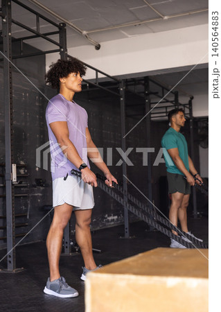 Diverse male gym partners gripping battle ropes on gym rig while generating rope waves, copy space Diverse male gym partners gripping battle ropes on gym rig while generating rope waves, copy space 140564883