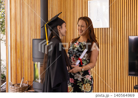 Asian mother-daughter standing in living room holding diploma wearing cap and gown Asian mother-daughter standing in living room holding diploma wearing cap and gown 140564969