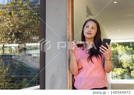 Asian woman standing near doorframe at home holding clear glass teacup and smartphone Asian woman standing near doorframe at home holding clear glass teacup and smartphone 140564972