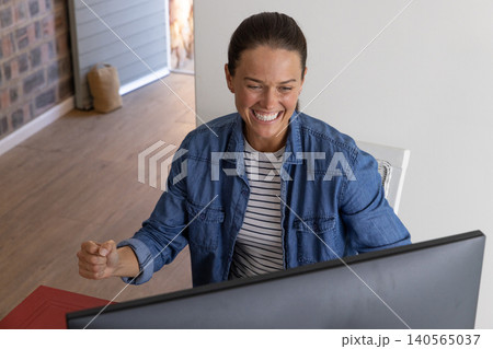 Woman leaning at desk in home office celebrating victory seen on large computer monitor 140565037