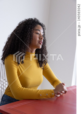 African american woman wearing mustard top sitting on wicker chair at table in room corner African american woman wearing mustard top sitting on wicker chair at table in room corner 140565042