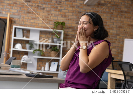 Asian woman covering mouth at office desk with computer and mug, copy space 140565057