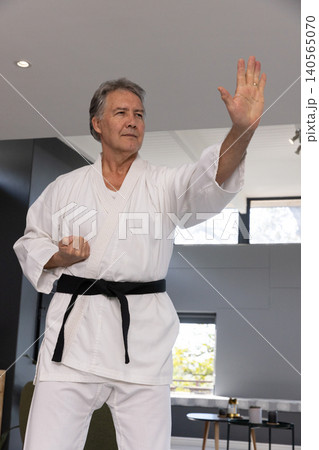 Senior male in white karate gi and belt demonstrating block stance in room with window Senior male in white karate gi and belt demonstrating block stance in room with window 140565070