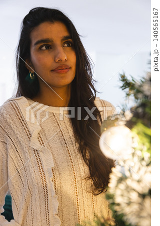 Asian female gazing to side in white room with green garland, glowing bulb and round ornament Asian female gazing to side in white room with green garland, glowing bulb and round ornament 140565167