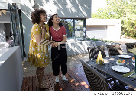 African american and asian female friends laughing and holding hands on deck, dining table African american and asian female friends laughing and holding hands on deck, dining table 140565179