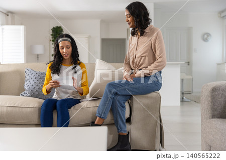Reading printed letter, diverse mom and teenage daughter sitting on sofa at home Reading printed letter, diverse mom and teenage daughter sitting on sofa at home 140565222