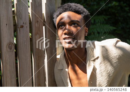 African american man leaning against wooden fence among foliage in sunlit backyard in shirt African american man leaning against wooden fence among foliage in sunlit backyard in shirt 140565248