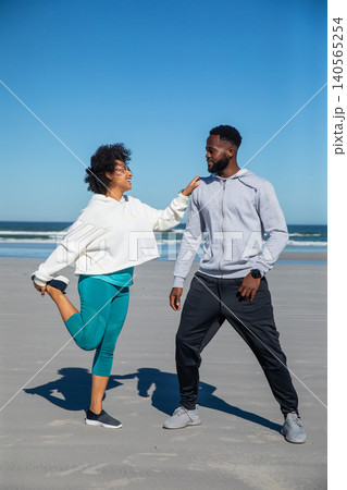 Diverse couple stretching quads on sandy beach wearing hoodies, leggings, sneakers, smartwatch 140565254