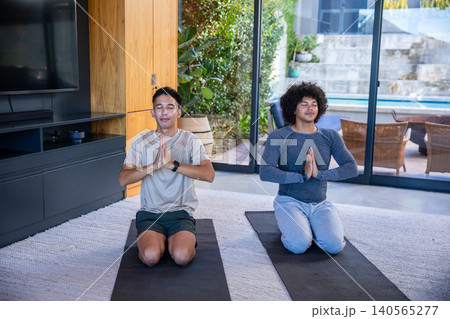 Diverse male friends practicing meditation kneeling on yoga mats at home facing patio chairs, pool Diverse male friends practicing meditation kneeling on yoga mats at home facing patio chairs, pool 140565277