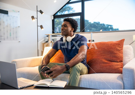 African American man sitting on sofa in living room using smartphone typing on laptop African American man sitting on sofa in living room using smartphone typing on laptop 140565296