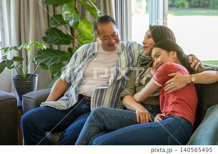 Diverse family sitting on gray sofa at home with plants parents holding daughter wearing wristwatch Diverse family sitting on gray sofa at home with plants parents holding daughter wearing wristwatch 140565495