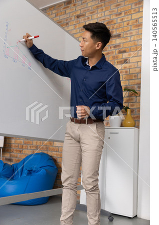 Adult Asian man wearing navy shirt standing drawing chart on whiteboard at office with red marker Adult Asian man wearing navy shirt standing drawing chart on whiteboard at office with red marker 140565513
