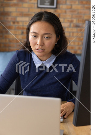 Adult Asian woman wearing navy sweater working at desk in office, using laptop and vertical monitor Adult Asian woman wearing navy sweater working at desk in office, using laptop and vertical monitor 140565520