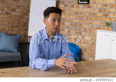 Asian male sitting at wooden table in office, wearing light blue shirt, gesturing near flip chart 140565521