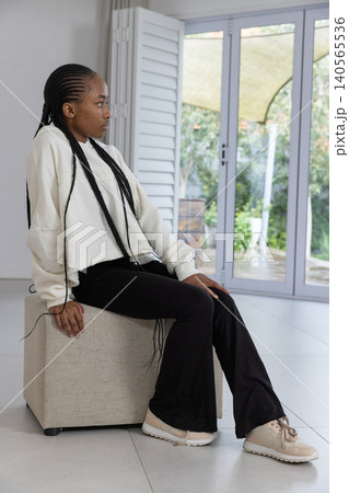 African American woman sitting on beige ottoman on tile floor near glass doors, wearing sweatshirt African American woman sitting on beige ottoman on tile floor near glass doors, wearing sweatshirt 140565536