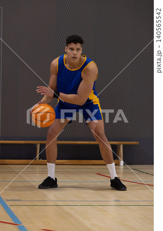 Male 20s in blue jersey holding orange basketball lowering into wide stance on court by bench 140565542
