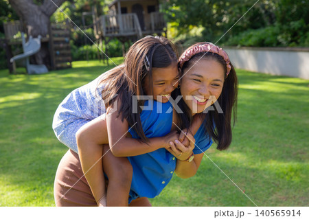 Mother and daughter giving piggyback ride on green lawn in backyard next to wooden treehouse Mother and daughter giving piggyback ride on green lawn in backyard next to wooden treehouse 140565914