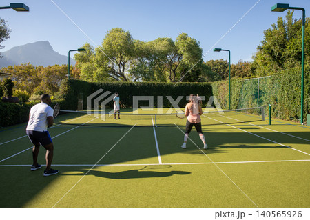 Diverse friends playing doubles tennis with rackets by net and hedges on backyard court Diverse friends playing doubles tennis with rackets by net and hedges on backyard court 140565926