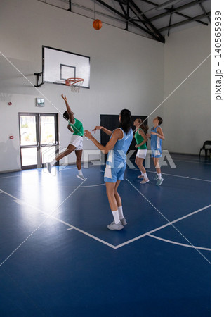 Diverse female basketball players jumping and reaching for basketball near hoop on blue court Diverse female basketball players jumping and reaching for basketball near hoop on blue court 140565939
