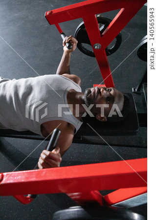 Mid adult African American man lying on bench pushing red chest-press machine weights at gym 140565943