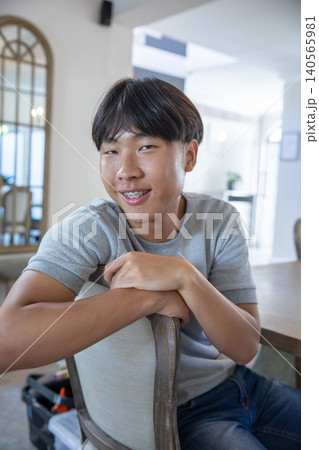 Teenage asian male leaning forward on wooden chair in residential dining room next to arched mirror 140565981