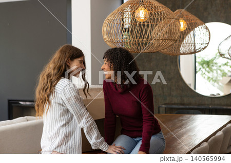 Female partners leaning on wooden dining table at home, smiling across moving box, mirror, lamps 140565994