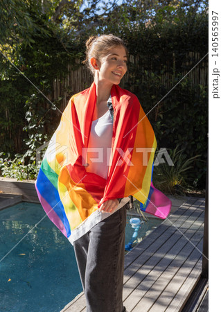 Female adult standing on backyard pool deck draped in rainbow pride flag under sunlight 140565997