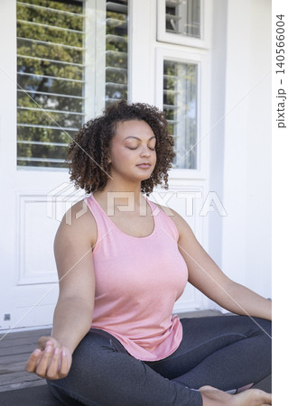 Adult African American woman in pink tank top leggings meditating on porch by door shuttered window Adult African American woman in pink tank top leggings meditating on porch by door shuttered window 140566004