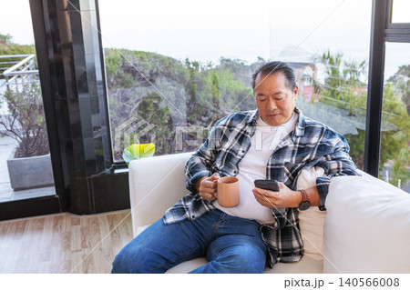 Asian man sipping from ceramic mug, scrolling smartphone on sofa in living room, copy space Asian man sipping from ceramic mug, scrolling smartphone on sofa in living room, copy space 140566008