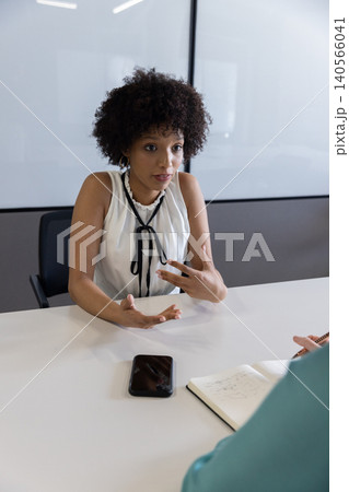 Female coworkers speaking and gesturing at conference table with smartphone, notebook and pen Female coworkers speaking and gesturing at conference table with smartphone, notebook and pen 140566041