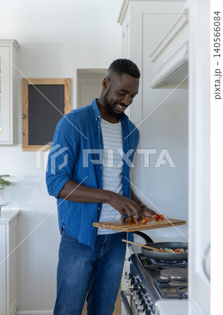 African american man slicing cherry tomatoes on cutting board by gas stove in home kitchen 140566084