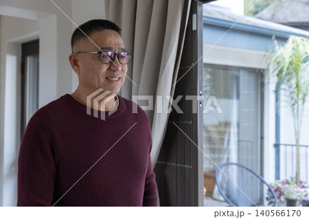 Senior chinese man looking out sliding glass door to patio at home with curtains 140566170
