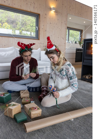 Couple sitting on gray rug in living room wrapping gifts with scissors, ribbon and kraft paper Couple sitting on gray rug in living room wrapping gifts with scissors, ribbon and kraft paper 140566171