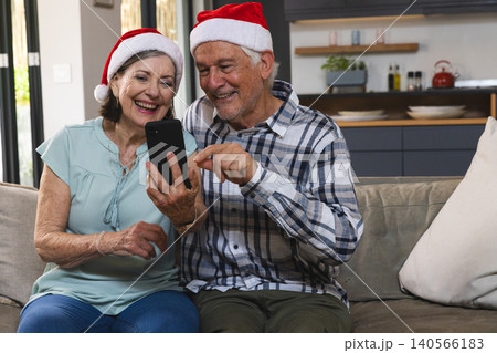 Senior couple sitting on beige couch in living room wearing Santa hats and viewing smartphone Senior couple sitting on beige couch in living room wearing Santa hats and viewing smartphone 140566183